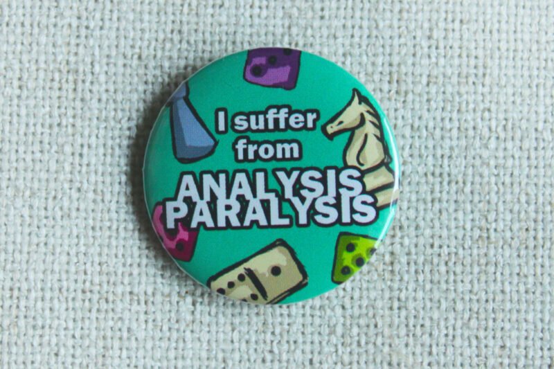 A seafoam green coloured button badge showing dice, a domino, a classic pawn and a knight chess piece and the text: I suffer from Analysis Paralysis.