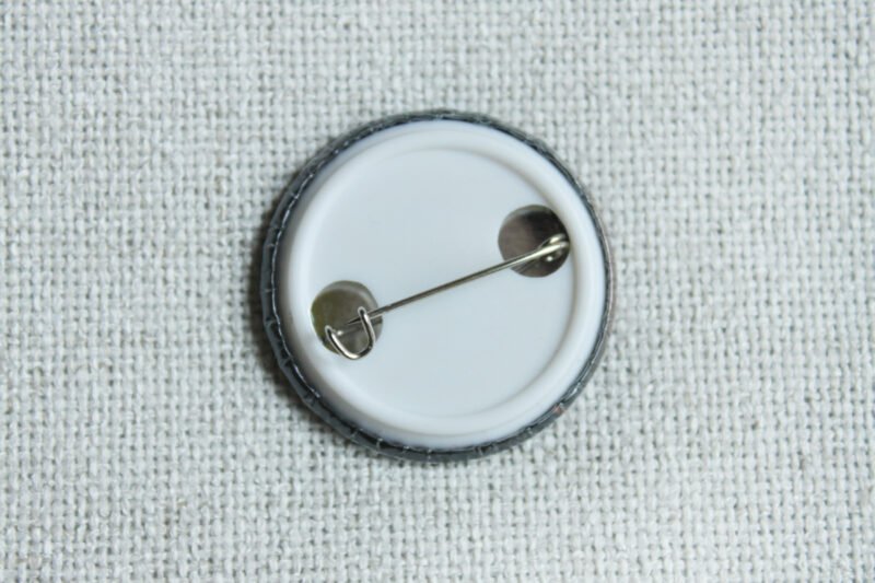 The white coloured back of a button badge with a dark coloured edge lying on cream coloured textile.