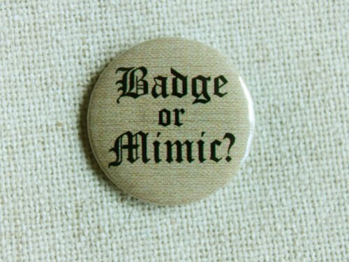 A beige coloured button badge showing the text “Badge or Mimic” in a medieval Blackletter font sitting on cream coloured textile.