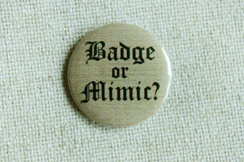 A beige coloured button badge showing the text “Badge or Mimic” in a medieval Blackletter font sitting on cream coloured textile.