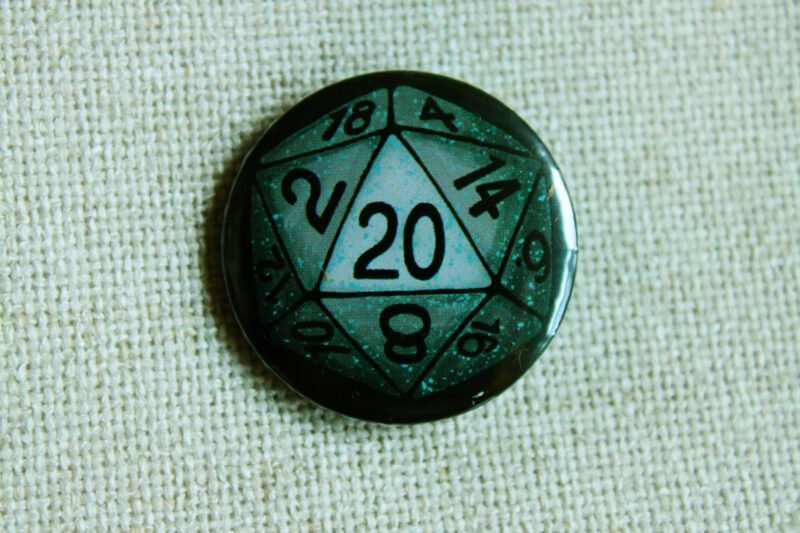 A black coloured button badge showing an illustrated blue and grey 20 sided die on the number 20 side.