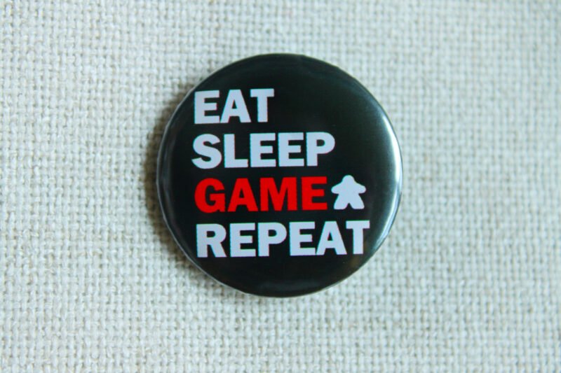 A black button badge showing the text Eat Sleep Game Repeat and the Meeple Silhouette. The word game is in red, the rest in white.