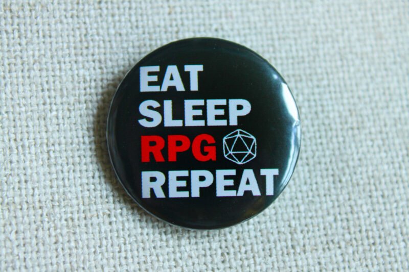 A black button badge showing the text Eat Sleep RPG Repeat and a D20 outline. The word RPG is in red, the rest is in white.