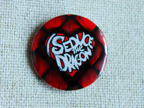 A button badge showing red reptilian scales and the text I Seduce the Dragon across a classic shaped heart.