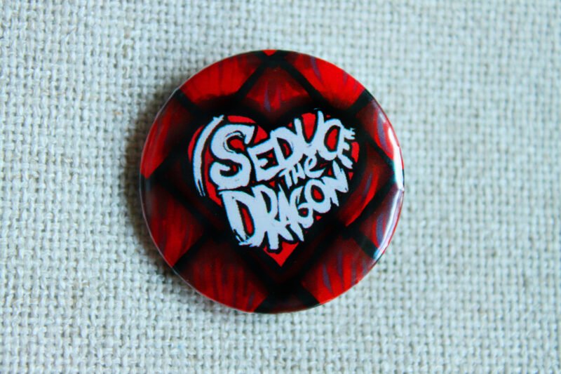 A button badge showing red reptilian scales and the text I Seduce the Dragon across a classic shaped heart.