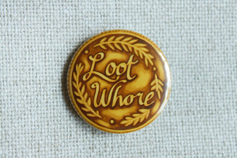 A gold coloured button badge illustrated to look like a coin with the text Loot Whore on it and decorative leaves.