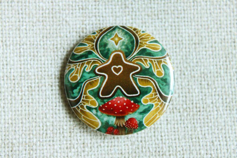 A button badge of a gingerbread meeple with faerie wings a star and fly agaric toadstools