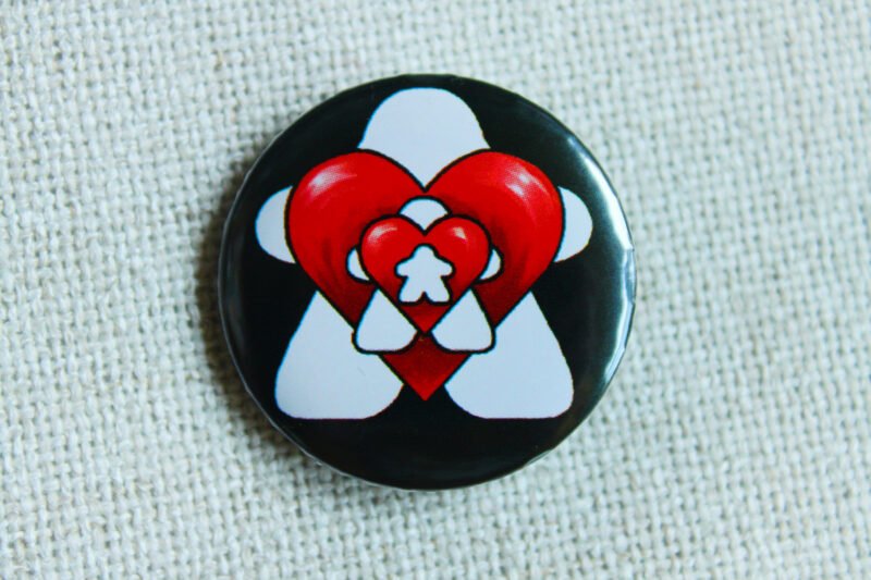 A black button badge whowing repeated images of white meeples and red hearts alternatively getting smaller.