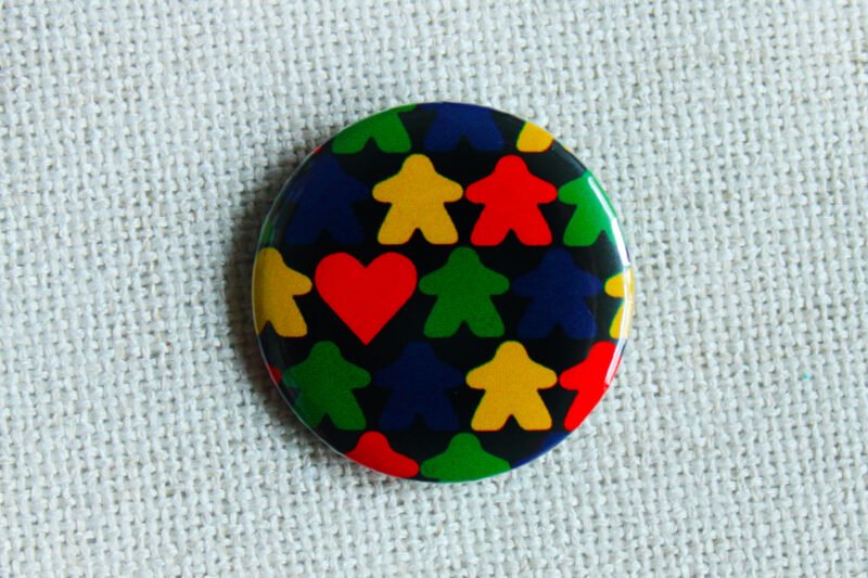 A black button badge with rows of meeple silhouettes in the colours red yellow green and blue and a classic shaped red heart.