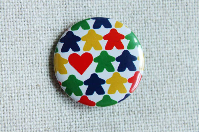 A white button badge with rows of meeple silhouettes in the colours red, yellow, green and blue and a classic shaped red heart.