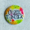 A button badge in yellows, greens, oranges and pinks with black specs and the hand drawn text Overthinker wherein each letter is a different font.