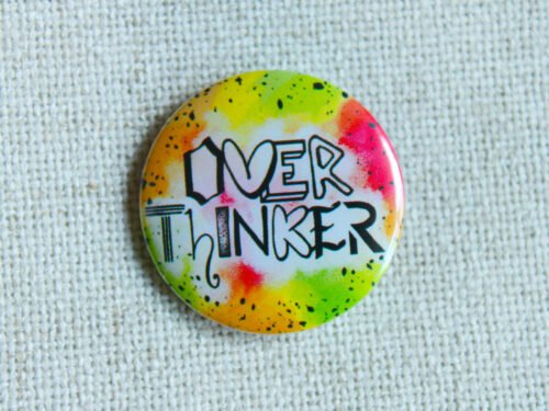 A button badge in yellows, greens, oranges and pinks with black specs and the hand drawn text Overthinker wherein each letter is a different font.