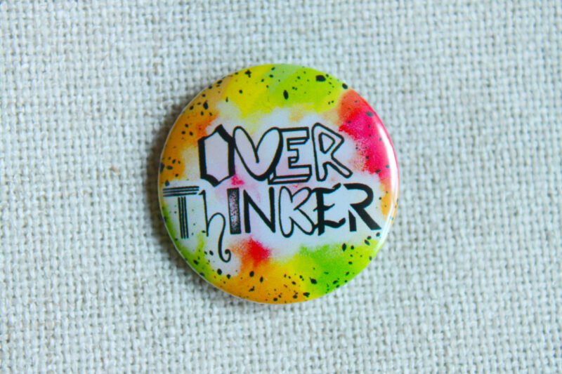 A button badge in yellows, greens, oranges and pinks with black specs and the hand drawn text Overthinker wherein each letter is a different font.