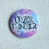 A button badge in purples, blues and pinks with black specs and the hand drawn text Overthinker wherein each letter is a different font.