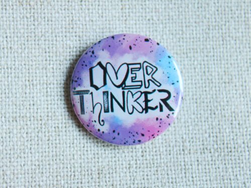 A button badge in purples, blues and pinks with black specs and the hand drawn text Overthinker wherein each letter is a different font.