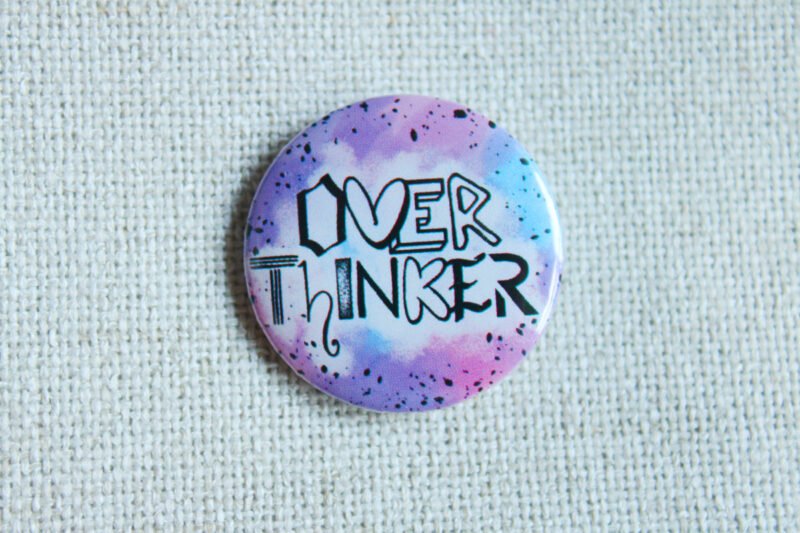 A button badge in purples, blues and pinks with black specs and the hand drawn text Overthinker wherein each letter is a different font.