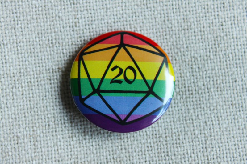 A button badge with a rainbow striped background and the outline of a D20 die in black.