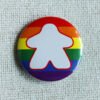 A button badge with a rainbow striped background and the white silhouette of a meeple with a red outline.