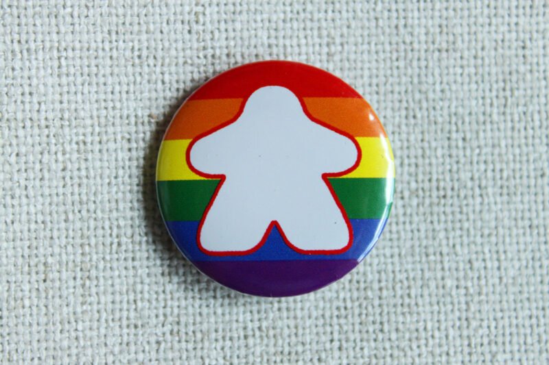 A button badge with a rainbow striped background and the white silhouette of a meeple with a red outline.