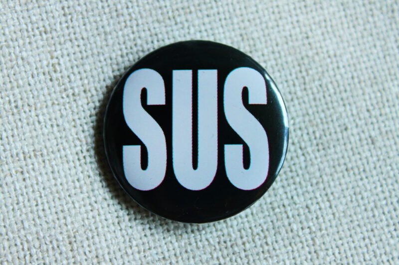 A black button badge showing the large white all caps word SUS as an abbreviation of Suspisious