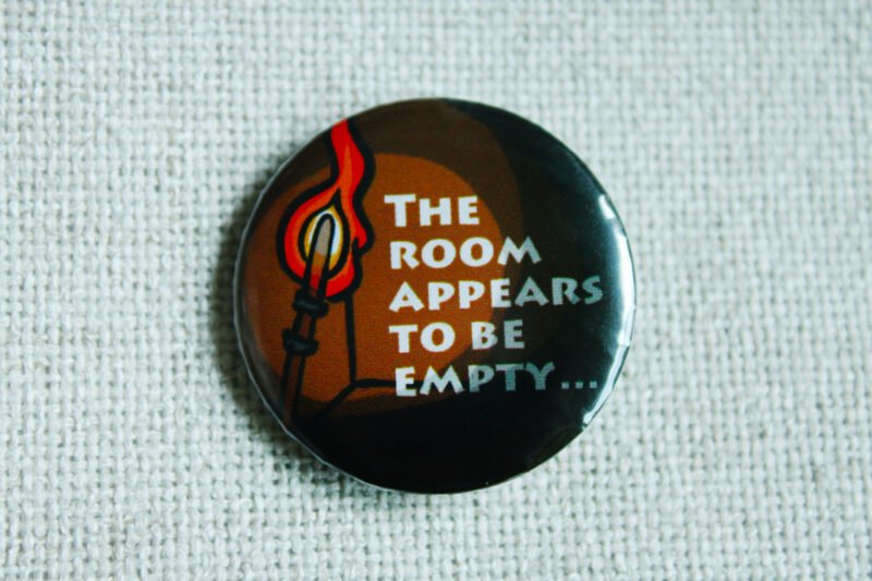 An illustrated button badge showing a fire torch and a dark empty room with the text The Room Appears to be Empty