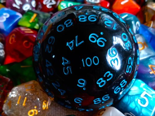 A large 100-sided die in black with blue numbers resting on a bed of colourful polyhedral dice.