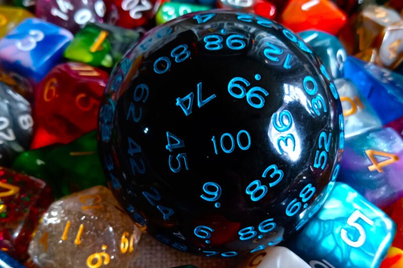 A large 100-sided die in black with blue numbers resting on a bed of colourful polyhedral dice.