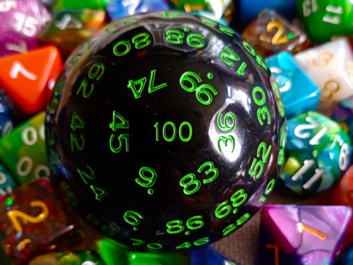 A large 100-sided die in black with green numbers resting on a bed of colourful polyhedral dice.