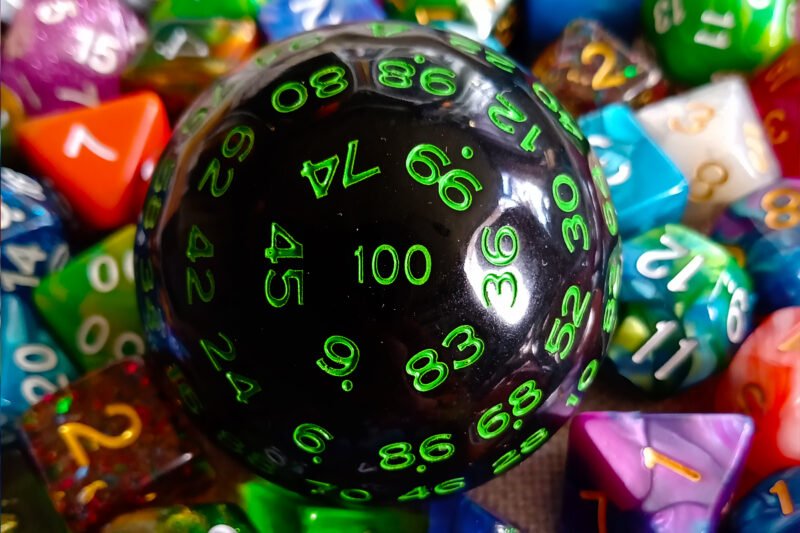 A large 100-sided die in black with green numbers resting on a bed of colourful polyhedral dice.