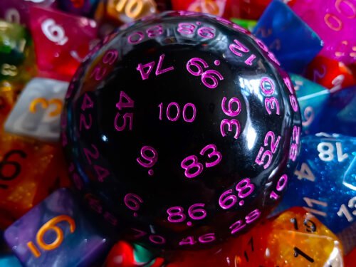 A large 100-sided die in black with purple numbers resting on a bed of colourful polyhedral dice.