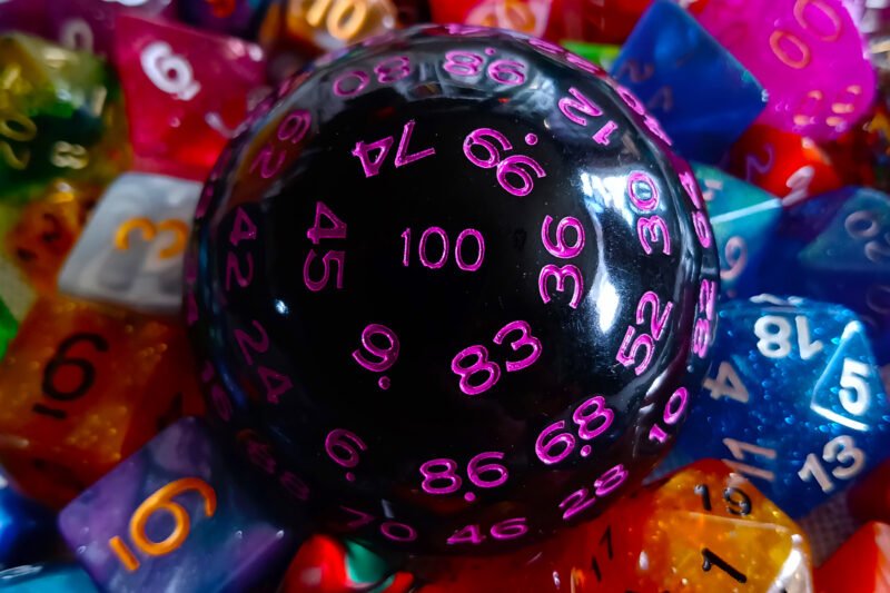 A large 100-sided die in black with purple numbers resting on a bed of colourful polyhedral dice.