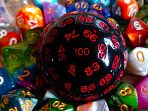 A large 100-sided die in black with red numbers resting on a bed of colourful polyhedral dice.