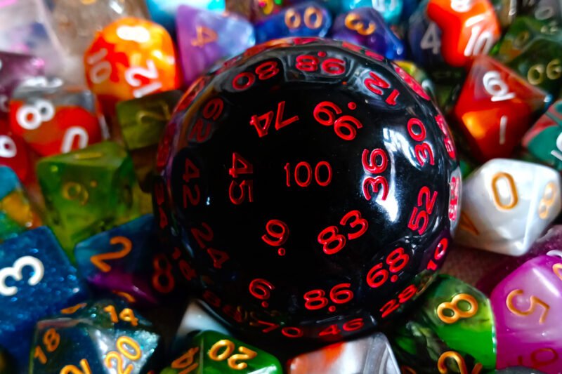 A large 100-sided die in black with red numbers resting on a bed of colourful polyhedral dice.