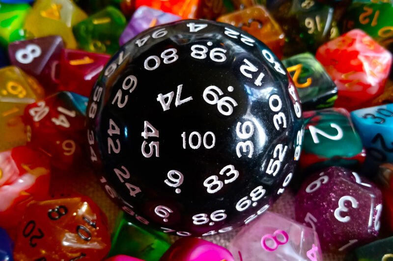 A large 100-sided die in black with white numbers resting on a bed of colourful polyhedral dice.