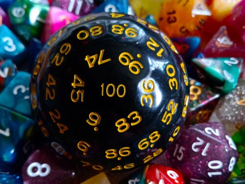 A large 100-sided die in black with yellow numbers resting on a bed of colourful polyhedral dice.