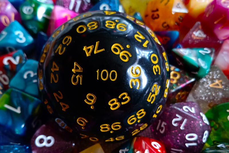 A large 100-sided die in black with yellow numbers resting on a bed of colourful polyhedral dice.