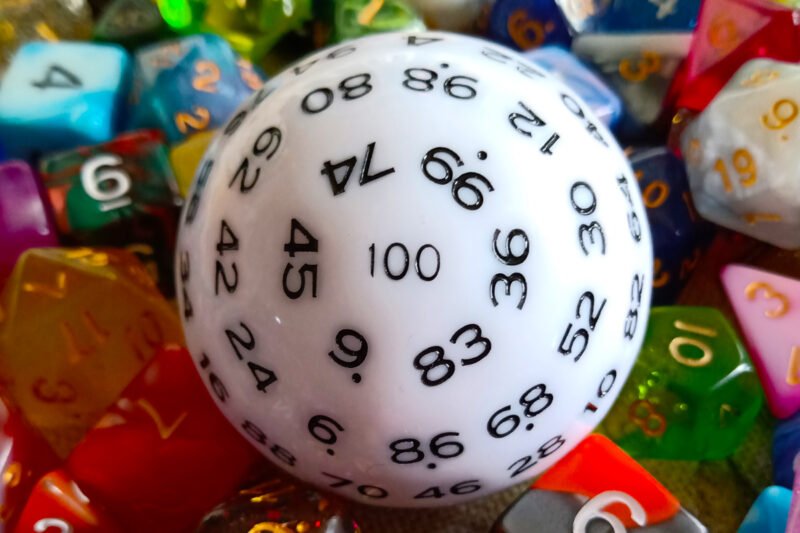 A large 100-sided die in white with black numbers resting on a bed of colourful polyhedral dice.