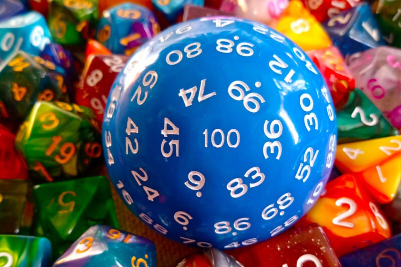 A large 100-sided die in blue with white numbers resting on a bed of colourful polyhedral dice.