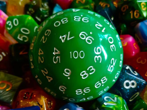 A large 100-sided die in green with white numbers resting on a bed of colourful polyhedral dice.