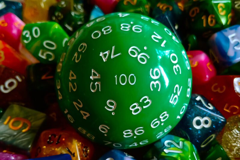 A large 100-sided die in green with white numbers resting on a bed of colourful polyhedral dice.