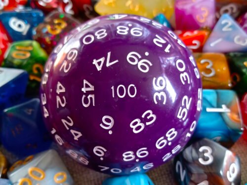 A large 100-sided die in purple with white numbers resting on a bed of colourful polyhedral dice.