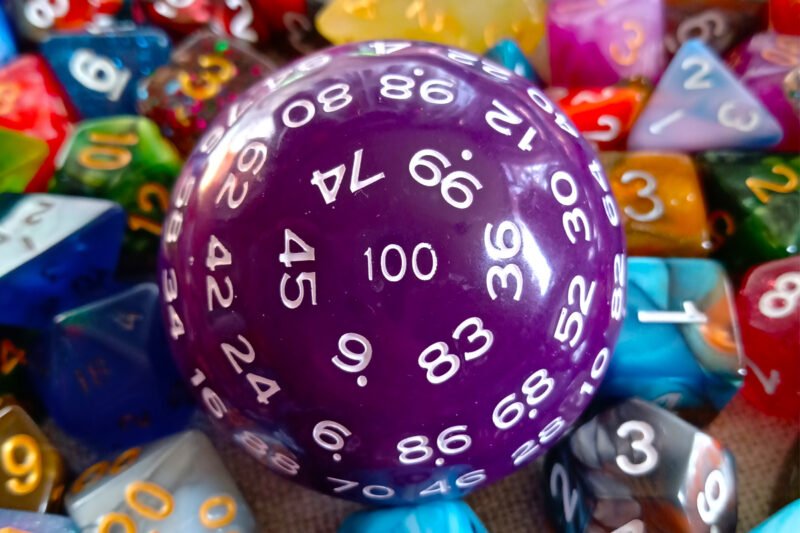 A large 100-sided die in purple with white numbers resting on a bed of colourful polyhedral dice.