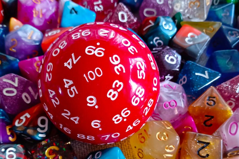 A large 100-sided die in red with white numbers resting on a bed of colourful polyhedral dice.