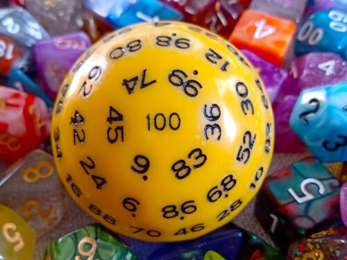 A large 100-sided die in yellow with black numbers resting on a bed of colourful polyhedral dice.