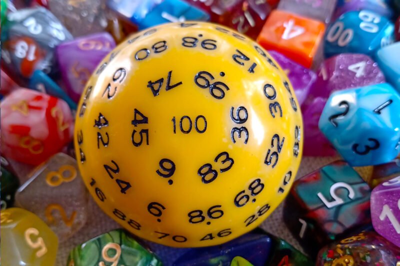 A large 100-sided die in yellow with black numbers resting on a bed of colourful polyhedral dice.