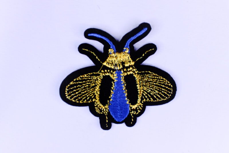 An embroidered patch of a winged bug in the colours black gold and blue.