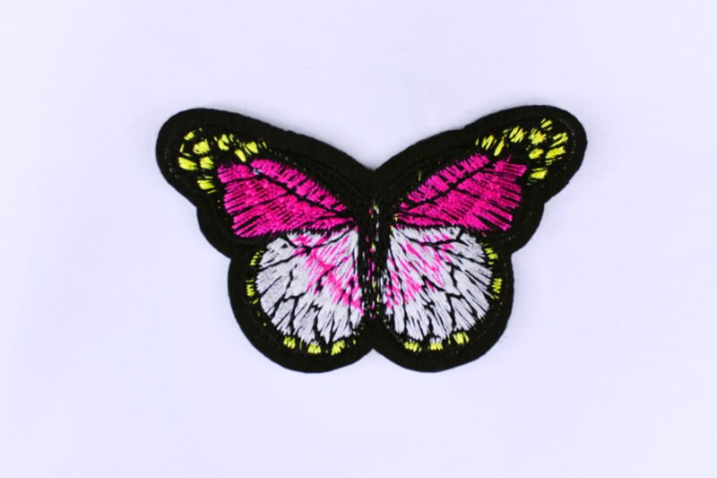 An embroidered patch of a butterfly in the style of a monarch butterfly but in the colours black white pink and yellow.