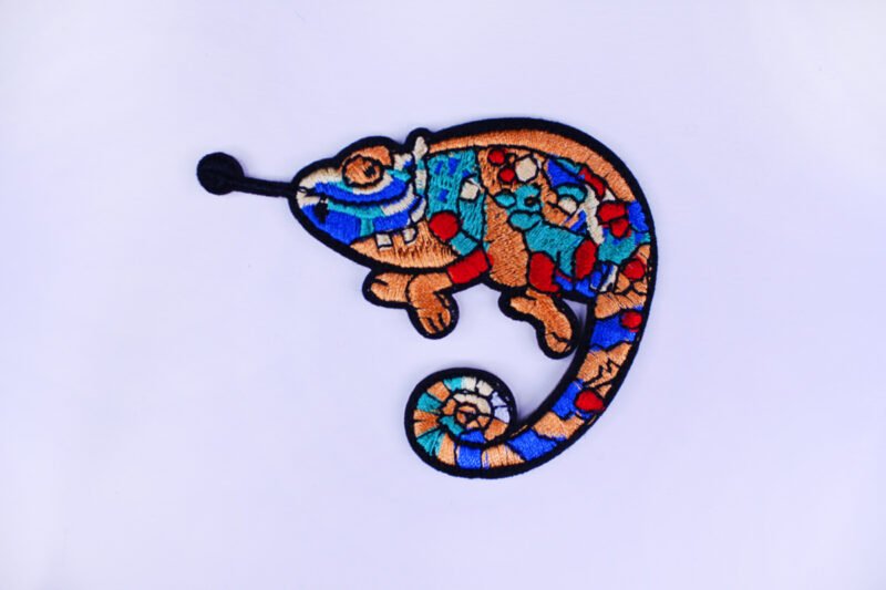 An embroidered patch of a chameleon with colourful scales and a curled up tail and poking out its tongue.