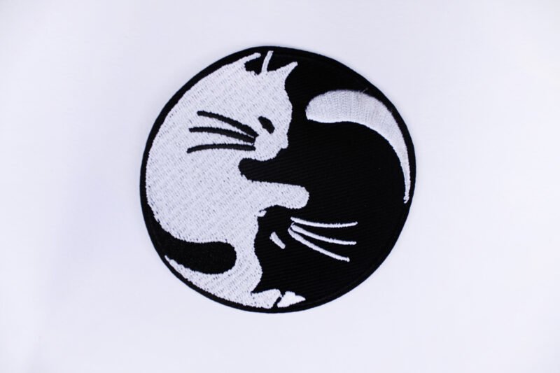 An embroidered patch of a ying yang symbol made up of a black and a white cat cuddling.