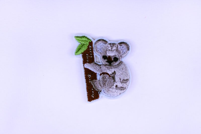 An embroidered patch of a small grey koala hanging onto a branch with two green leaves on it.